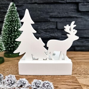 Könnte beinhalten: Weiße Holz-Weihnachtsdekoration mit einem Reh und zwei Tannenbäumen. Die Dekoration steht auf einem weißen Sockel.