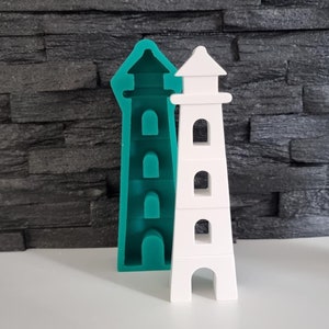 Könnte beinhalten: Eine weiße Leuchtturm-förmige Form mit einer blaugrünen Leuchtturm-Form dahinter. Beide Formen haben ein detailliertes Design mit Fenstern und einem spitzen Dach.