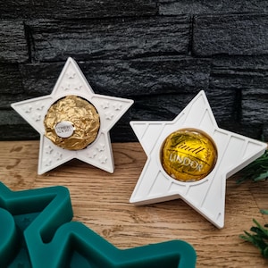 Könnte beinhalten: Zwei weiße sternförmige Schokoladenhalter mit einer goldenen Schokoladenbonbon in jedem. Der Halter links hat eine Ferrero Rocher Schokolade und der Halter rechts hat eine Lindor Schokolade.