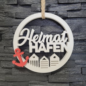 Könnte beinhalten: Weißer Holz-Kreis mit dem Text "Heimat Hafen" in einer dekorativen Schrift. Der Kreis ist mit einem roten Anker und drei weißen Strandhütten verziert.