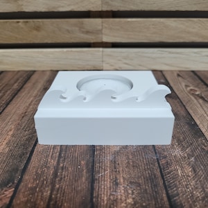 Könnte beinhalten: Ein weißer, rechteckiger Kerzenhalter mit einem Wellenmuster am oberen Rand. Der Halter hat eine kreisförmige Vertiefung für die Kerze. Der Kerzenhalter besteht aus einem festen, undurchsichtigen Material.