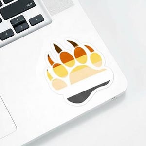May include: A sticker featuring a bear paw design with a color gradient of brown, orange, yellow, and white. The sticker is placed on a white laptop, with the keyboard visible in the background. The paw print has a black and gray stripe at the bottom.