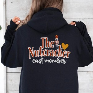 Puede incluir: Sudadera con capucha azul marino con el texto "The Nutcracker cast member" en escritura roja y blanca. Una pequeña ilustración de un cascanueces y un corazón dorado están encima del texto. La sudadera tiene capucha y mangas largas.