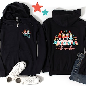 Puede incluir: Sudadera negra con cremallera y capucha con un diseño de Cascanueces en la parte posterior. La parte delantera de la sudadera tiene un diseño de Cascanueces más pequeño y el nombre "Molly" impreso en ella.