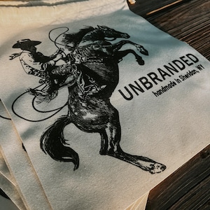 May include: Off-white canvas drawstring bags with a black illustration of a cowboy riding a rearing horse. The bags are printed with the word "UNBRANDED" and "handmade in Sheridan, WY".