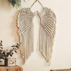 Op de afbeelding: Crèmekleurige macramé engelenvleugels wanddecoratie. De vleugels zijn gemaakt van geweven katoenen touw, met een gedetailleerd verenpatroon. Ze worden met een metalen haak aan de muur gehangen. Een decoratief item.