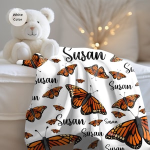 May include: White fleece blanket with a repeating pattern of orange and black monarch butterflies and the name "Susan" printed in black.