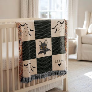 May include: A beige and black checkered baby blanket with a bat theme. The blanket features a cute bat illustration in the center square, bat silhouettes, and the name "LIAM" with a heart. The blanket is draped over a white crib.
