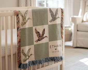May include: A woven baby blanket with a duck pattern in shades of green, brown, and beige, draped over a white crib. The blanket features a checkered design with ducks in flight. The name "ETHAN" and a date are displayed on the blanket.