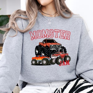 May include: Gray sweatshirt with the text "MOMSTER JAM" in red. The sweatshirt features a cartoon illustration of three monster trucks, one large and two smaller, with flames.