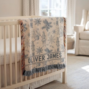 May include: A woven throw blanket featuring a blue and beige pattern of nutcrackers, snowflakes, and pine branches. The blanket includes the name OLIVER JAMES printed in a dark blue font near the bottom edge and has a fringed border.