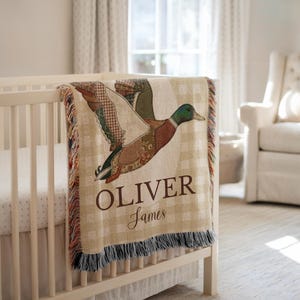 May include: A beige and brown baby blanket with a duck design and the name "OLIVER" printed on it. The blanket is draped over a white crib. The duck has green, brown, and white patches. The blanket has a fringed edge.
