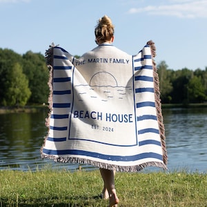 May include: A cream-colored beach blanket with blue stripes and fringe, held by a person standing near a lake. The blanket features the text "THE MARTIN FAMILY BEACH HOUSE EST. 2024" in a navy blue font, with a sun and water design.