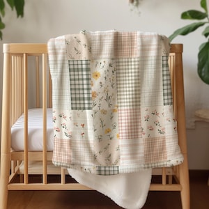 May include: A patchwork baby blanket with floral and plaid patterns in soft pastel colors, draped over a wooden crib. The blanket features a white, fluffy underside, creating a cozy and inviting aesthetic.