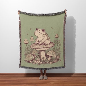 May include: A green woven throw blanket with a cartoon illustration of a frog sitting on a mushroom. The mushroom has brown spots and is surrounded by other mushrooms and green leaves.