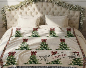May include: A cream-colored Christmas throw blanket featuring green Christmas trees adorned with red bows and white lights. The blanket has a black fringe and a banner that reads "Johnsons".