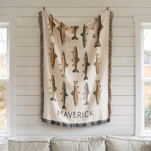 May include: Cream-colored woven throw blanket featuring illustrations of various fish in shades of brown, green, and gray. The word "MAVERICK" is printed in black at the bottom. The blanket has a fringed edge.