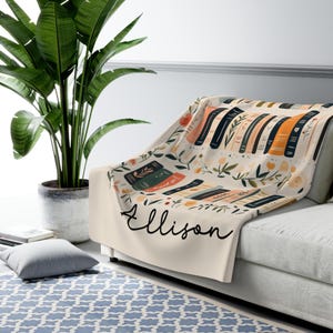 May include: A cream-colored fleece throw blanket with a floral pattern and stacks of books. The blanket has the name "Ellison" written in black cursive.