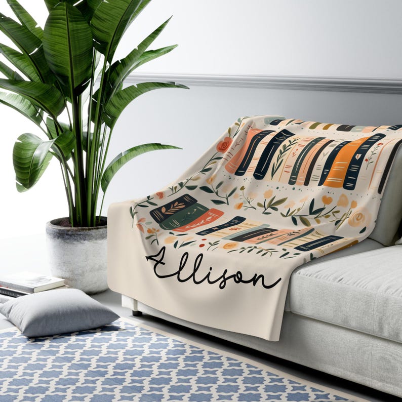 May include: A cream-colored fleece throw blanket with a floral pattern and stacks of books. The blanket has the name "Ellison" written in black cursive.