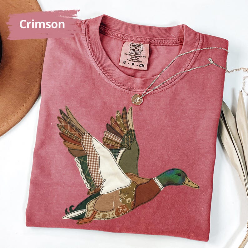 K&ouml;nnte beinhalten: Karmesinrotes T-Shirt mit einem Patchwork-Design einer fliegenden Ente. Die Ente hat einen gr&uuml;nen Kopf, einen braunen K&ouml;rper und Fl&uuml;gel aus verschiedenen gemusterten Stoffen. Eine silberne Halskette mit einem kleinen Anh&auml;nger liegt auf dem Shirt.