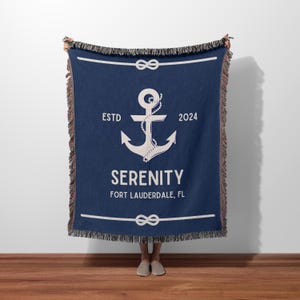 May include: A navy blue woven throw blanket featuring a large white anchor graphic wrapped in rope. The text reads ESTD 2024, SERENITY, and FORT LAUDERDALE, FL. The blanket has a decorative fringe on the top and bottom edges and horizontal rope borders.