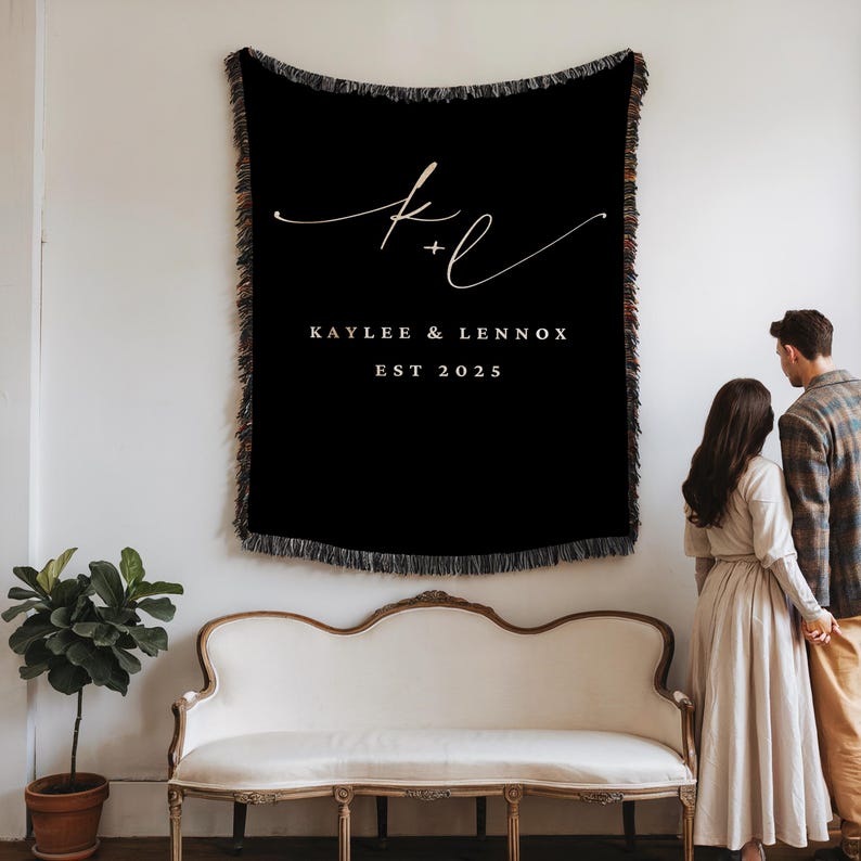 Op de afbeelding: Zwarte geweven plaid met cr&egrave;mekleurige franjes, met de namen "Kaylee & Lennox" en het jaar "EST 2025" in cr&egrave;mekleurig schrift. De plaid hangt aan een witte muur boven een witte bank.