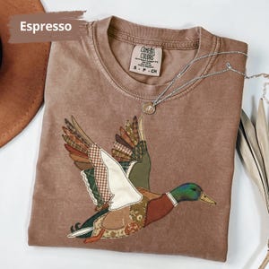K&ouml;nnte beinhalten: Espressofarbenes T-Shirt mit einem Patchwork-Enten-Design. Die Ente hat Fl&uuml;gel mit verschiedenen gemusterten Stoffen und einen gr&uuml;nen und braunen Kopf. Eine silberne Halskette mit Anh&auml;nger liegt &uuml;ber dem Shirt. Das Shirt ist mit "Comfort Colors" und Gr&ouml;&szlig;e S-CH gekennzeichnet.
