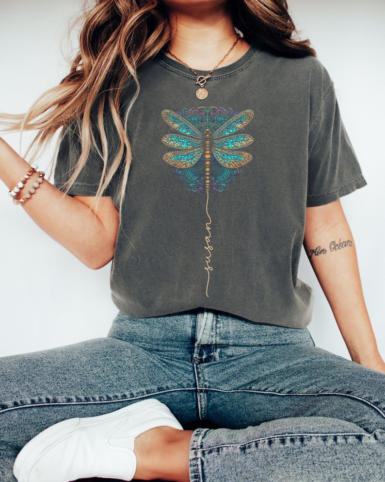 May include: A grey t-shirt with a blue and gold dragonfly design and the word "trust" written vertically down the centre of the shirt.