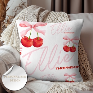 May include: A white pillow with a pink background featuring the name "Ellie Thompson" written in pink cursive. The pillow has a repeating pattern of pink bows and red cherries.