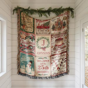 May include: A woven throw blanket featuring a grid of Christmas-themed illustrations and text, including a red truck, a gingerbread house, a snowman, and a central wreath with the letter J and the name Johnsons, finished with fringed edges.