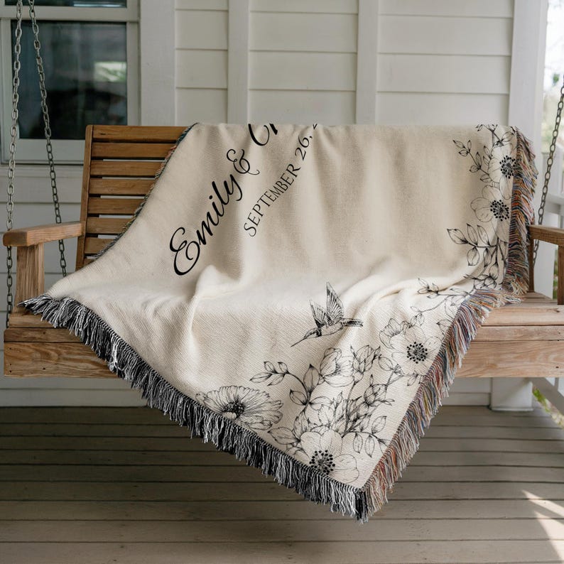 May include: Cream-colored woven throw blanket with black floral and hummingbird design. The blanket has a fringed edge and features the names "Emily & Chris" and the date "September 26, 20--" in black script.