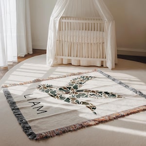 May include: A cream-colored baby blanket with a large duck graphic in shades of brown, green, and tan. The name "LIAM" is printed in black block letters. The blanket has a fringed edge and is placed on a white rug near a crib.