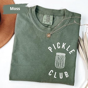 May include: A moss green t-shirt with the text "PICKLE CLUB" in white. The shirt features a white illustration of a jar of pickles.