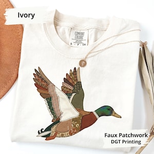 K&ouml;nnte beinhalten: Elfenbeinfarbenes T-Shirt mit einem Patchwork-Design eines fliegenden Entenmotivs. Die Ente hat Fl&uuml;gel mit verschiedenen gemusterten Stoffen in Braun-, Gr&uuml;n- und Beiget&ouml;nen. Das Shirt tr&auml;gt auch den Text "Faux Patchwork DGT Printing". Eine goldene Halskette mit Anh&auml;nger liegt auf dem Shirt.