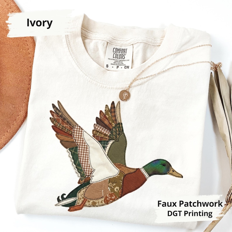 K&ouml;nnte beinhalten: Elfenbeinfarbenes T-Shirt mit einem Patchwork-Design eines fliegenden Entenmotivs. Die Ente hat Fl&uuml;gel mit verschiedenen gemusterten Stoffen in Braun-, Gr&uuml;n- und Beiget&ouml;nen. Das Shirt tr&auml;gt auch den Text "Faux Patchwork DGT Printing". Eine goldene Halskette mit Anh&auml;nger liegt auf dem Shirt.