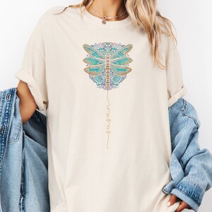 May include: A white t-shirt featuring a blue and gold dragonfly design with the word "surrender" written in gold script.