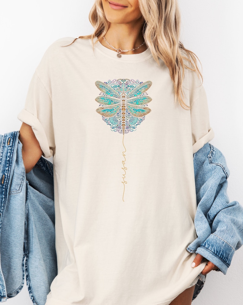 May include: A white t-shirt featuring a blue and gold dragonfly design with the word "surrender" written in gold script.