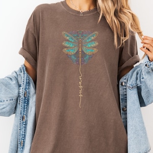 May include: An oversized brown t-shirt featuring a mandala-style dragonfly design and the word "inspire" written vertically in gold script.