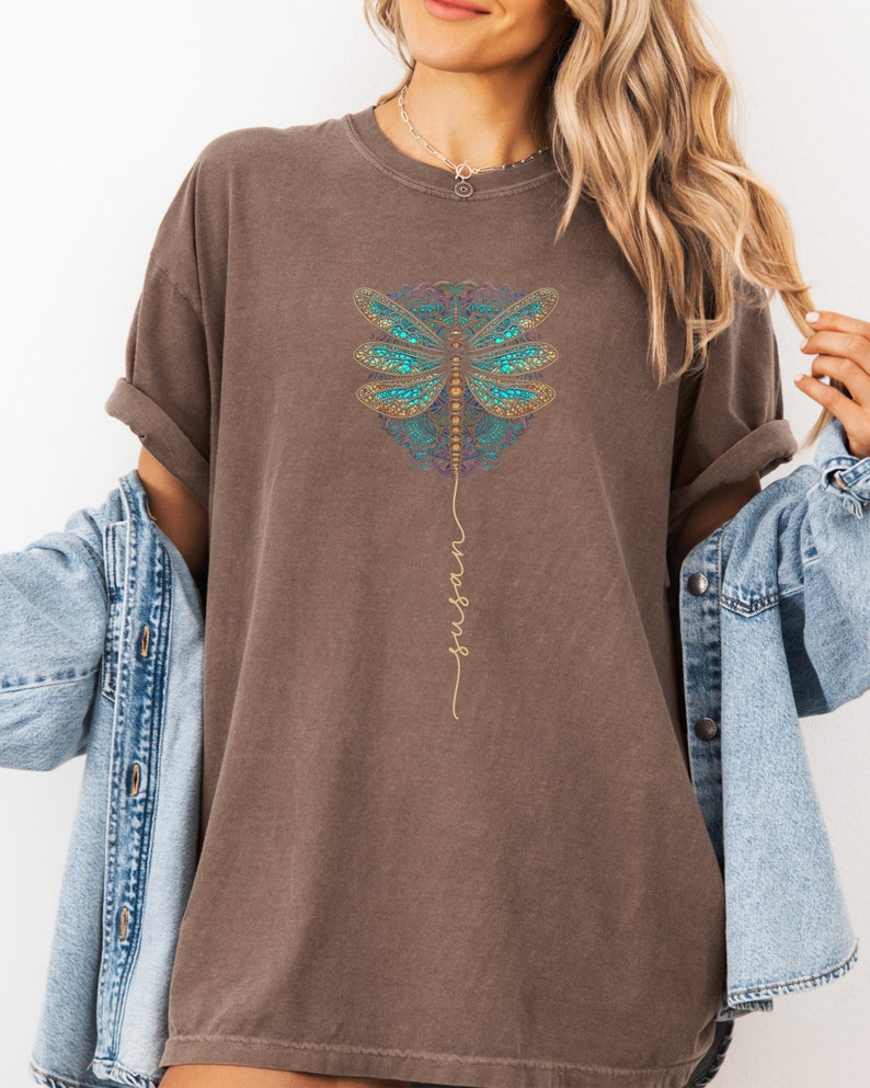 May include: An oversized brown t-shirt featuring a mandala-style dragonfly design and the word "inspire" written vertically in gold script.