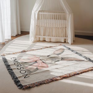 Puede incluir: Una manta rectangular para beb&eacute; con una ilustraci&oacute;n de acuarela de un conejito gris con un lazo rosa. El nombre "EMERSON" est&aacute; impreso en negro. La manta tiene un borde con flecos y est&aacute; sobre una alfombra blanca.