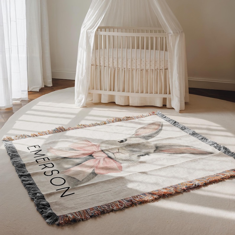 Puede incluir: Una manta rectangular para beb&eacute; con una ilustraci&oacute;n de acuarela de un conejito gris con un lazo rosa. El nombre "EMERSON" est&aacute; impreso en negro. La manta tiene un borde con flecos y est&aacute; sobre una alfombra blanca.