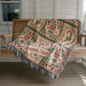 May include: A woven Christmas-themed throw blanket with a cream background and a repeating pattern of red and green holiday motifs. The blanket features a banner that reads "JOHNSON FAMILY". The blanket is draped over a wooden swing.