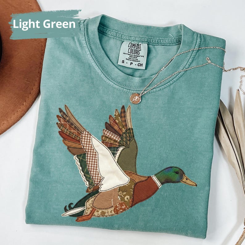 K&ouml;nnte beinhalten: Hellgr&uuml;nes T-Shirt mit einem Patchwork-Enten-Design im Flug. Die Ente hat braune, gr&uuml;ne und wei&szlig;e gemusterte Fl&uuml;gel und einen K&ouml;rper. Eine silberne Halskette mit einem kleinen Anh&auml;nger liegt auf dem Shirt. Der Text "Light Green" befindet sich oben links.