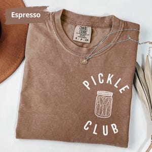 Peut inclure: Un t-shirt marron avec le texte "Pickle Club" en lettres blanches. Le t-shirt pr&eacute;sente un contour blanc d'un pot de cornichons.