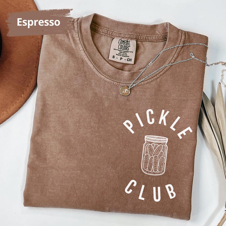 Peut inclure: Un t-shirt marron avec le texte "Pickle Club" en lettres blanches. Le t-shirt pr&eacute;sente un contour blanc d'un pot de cornichons.