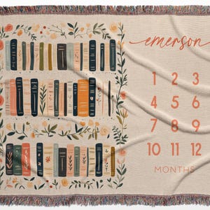 Op de afbeelding: Een beige baby mijlpaaldeken met een boekenplankontwerp en bloemaccenten. De deken heeft een boekenplankillustratie met verschillende boekkleuren en de naam "emerson" in cursief. Cijfers 1-12 worden weergegeven voor maandelijkse foto's.