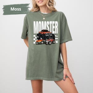 May include: A moss green t-shirt with the text "Momster Jam" in white. The shirt features a graphic of a red monster truck with black tyres and two smaller monster trucks.