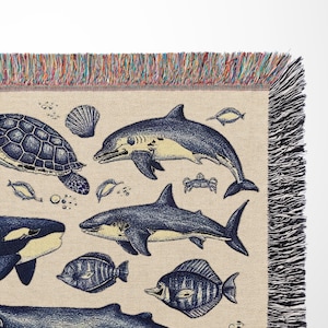 Puede incluir: Una manta beige con un dise&ntilde;o de vida marina. La manta presenta ilustraciones detalladas de varias criaturas marinas, incluyendo una tortuga, tiburones y peces, en tonos azules y amarillos. Los bordes est&aacute;n acabados con un borde con flecos.