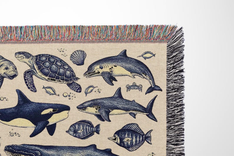 Puede incluir: Una manta beige con un dise&ntilde;o de vida marina. La manta presenta ilustraciones detalladas de varias criaturas marinas, incluyendo una tortuga, tiburones y peces, en tonos azules y amarillos. Los bordes est&aacute;n acabados con un borde con flecos.