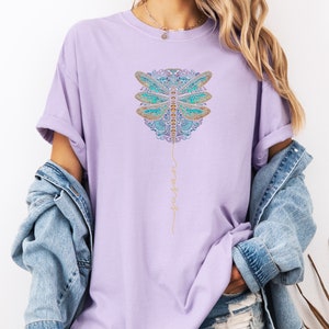 May include: A lavender t-shirt featuring a gold and blue dragonfly graphic with the word "trust" written vertically in gold script.